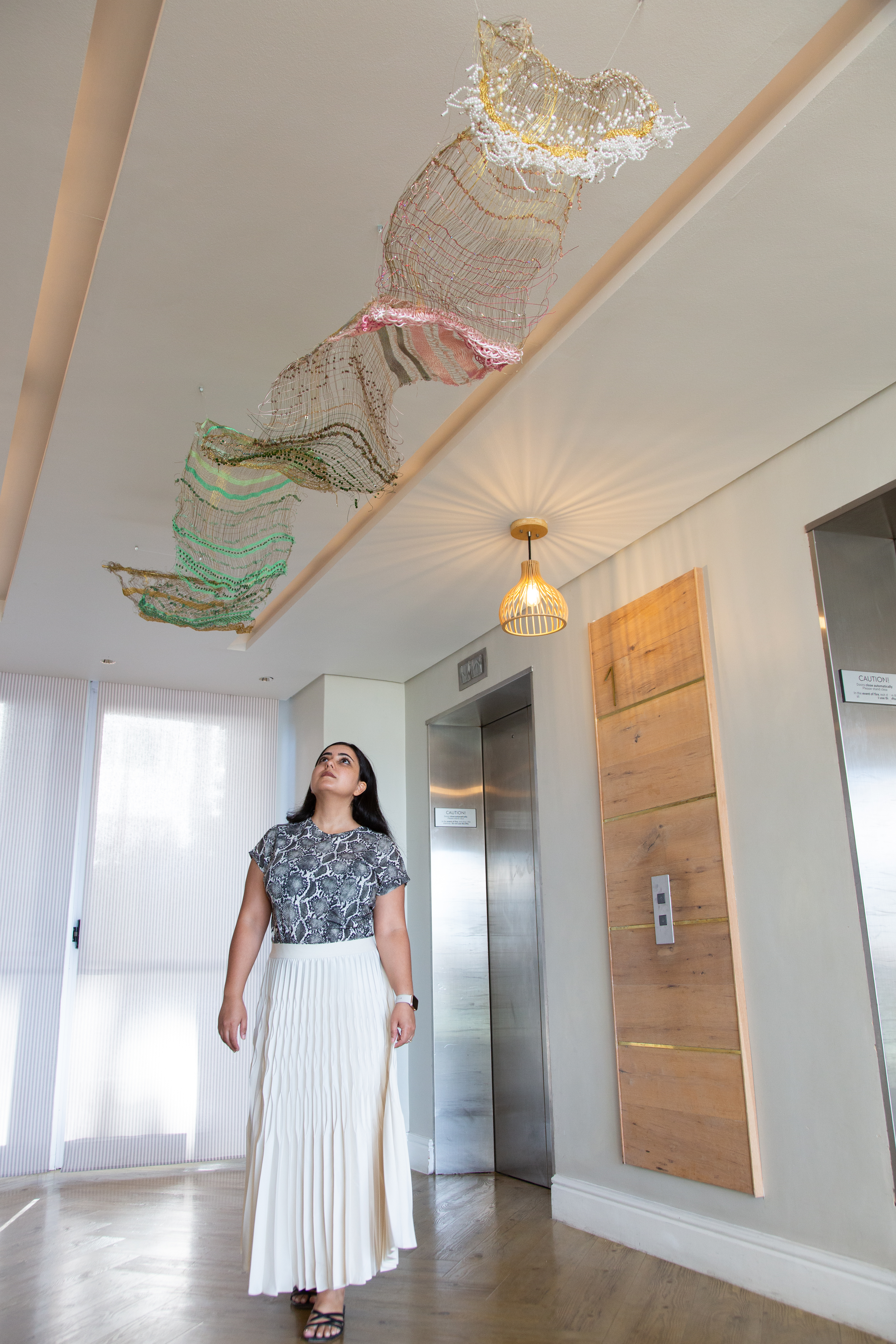 Sundus Saad standing beneath her large woven installation Jannah, suspended from the ceiling